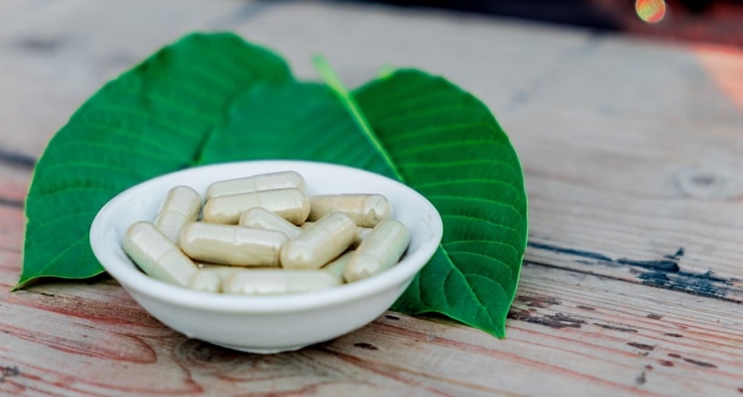 Kratom capsules in a bowl next to kratom leaves.
