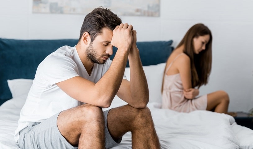Depressed man sitting on bed with girlfriend after quarrel about sexual problems.