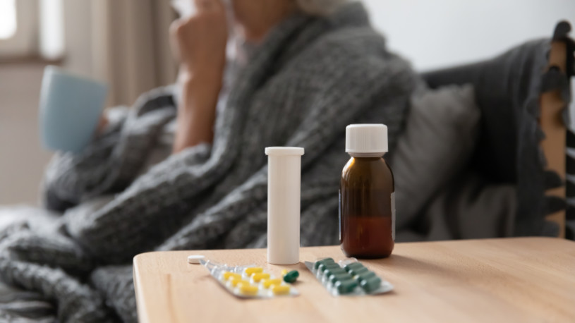 On bedside table blister pack with oral capsules syrup bottle close up, on background sick woman hold mug take antipyretic meds drink hot tea.