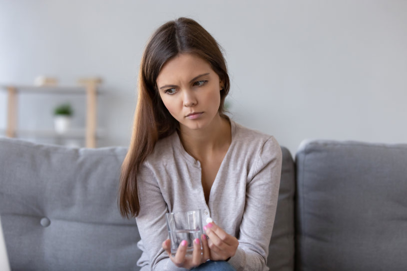 Lonely sad young woman sitting on sofa at home holding pill glass of water.