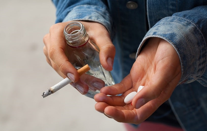 Woman smoking cigarettes and holding pills and vodka in her hands.