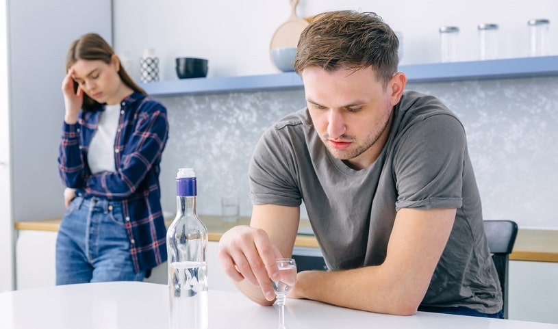 Man drinking alcohol and his wife standing behind depressed.
